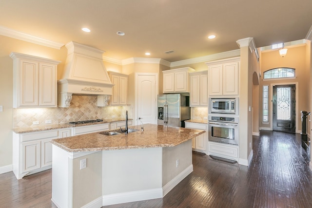kitchen featuring stainless steel appliances, a sink, premium range hood, dark wood-style flooring, and an island with sink
