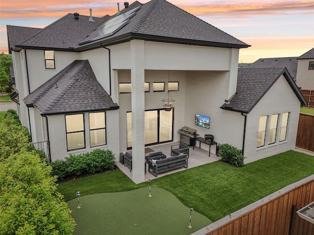 back of house at dusk with a shingled roof, an area to practice putting, a patio area, and stucco siding