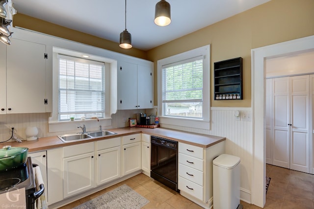 kitchen with black dishwasher, a wainscoted wall, a sink, light tile patterned floors, and white cabinets