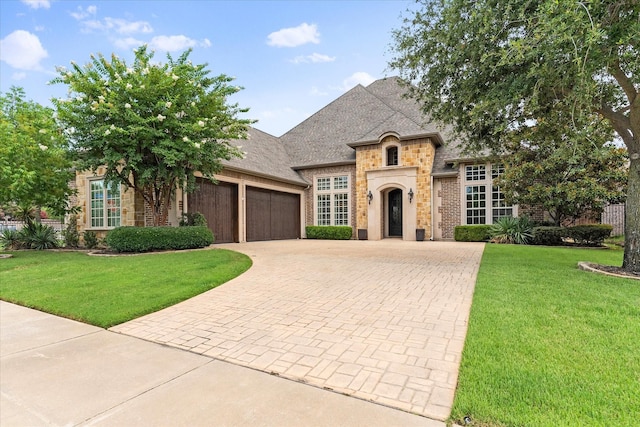 french provincial home featuring a garage, stone siding, decorative driveway, and a front lawn