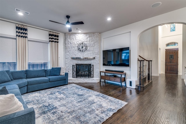 living room featuring arched walkways, a ceiling fan, a stone fireplace, wood finished floors, and stairs