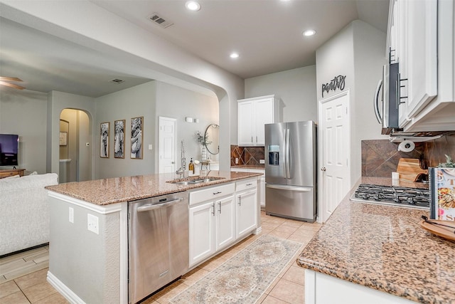 kitchen with stainless steel appliances, arched walkways, a sink, backsplash, and a center island with sink