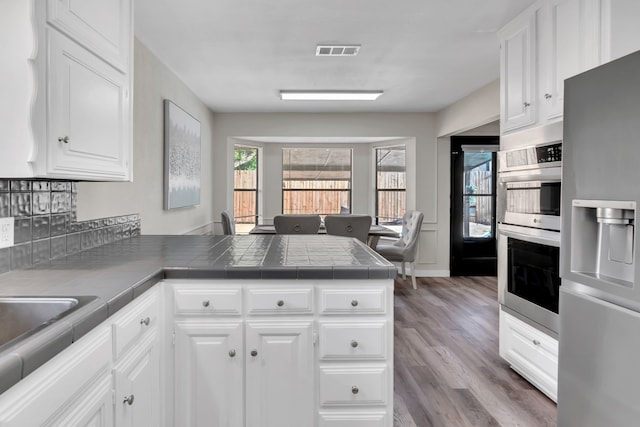 7108 Bermuda Ct, North Richland Hills, TX 76180 - kitchen featuring stainless steel appliances, a peninsula, wood finished floors, white cabinets, and tile countertops