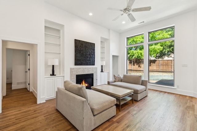 living room with built in shelves, a tiled fireplace, wood finished floors, ceiling fan, and baseboards