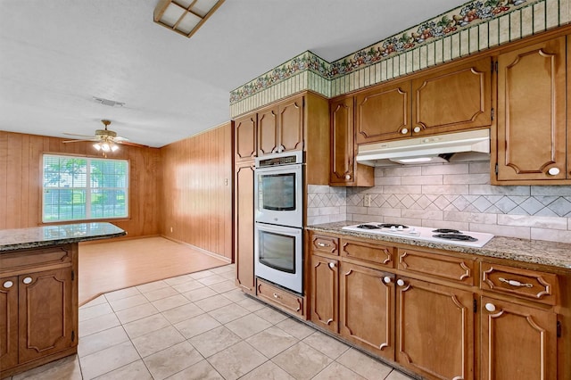 kitchen featuring under cabinet range hood, stainless steel double oven, white electric cooktop, brown cabinetry, and wooden walls