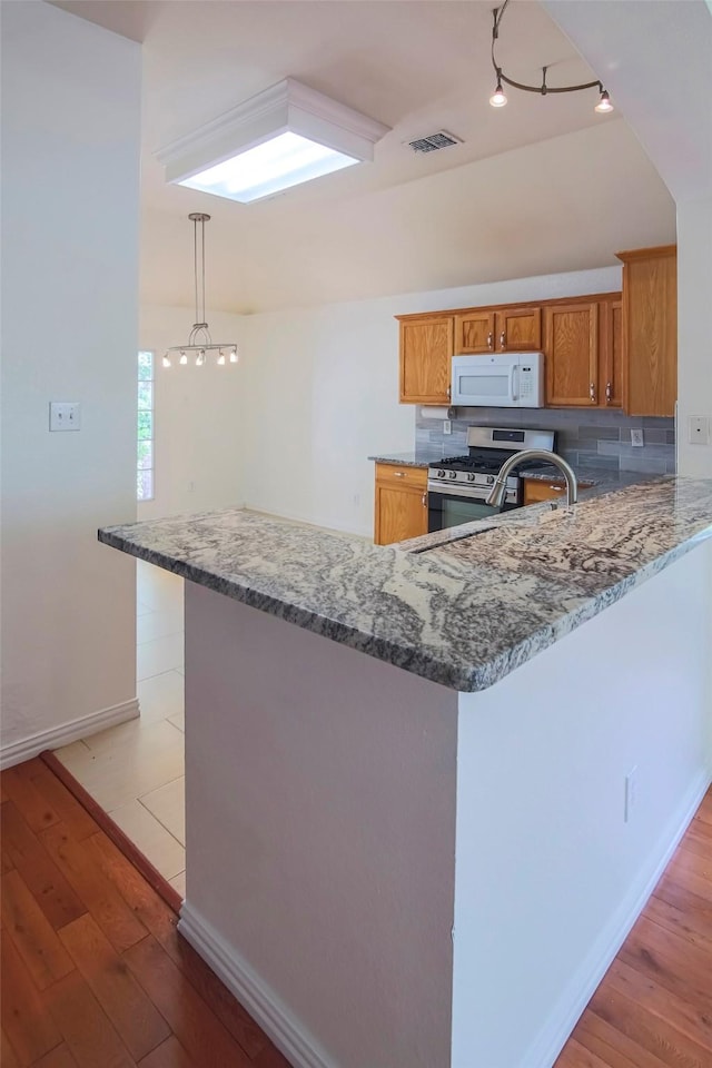 kitchen with white microwave, stainless steel range with gas stovetop, brown cabinetry, stone countertops, and light wood-style floors