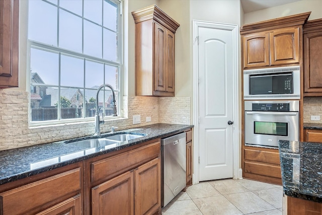 kitchen with appliances with stainless steel finishes, a sink, dark stone counters, tasteful backsplash, and light tile patterned floors