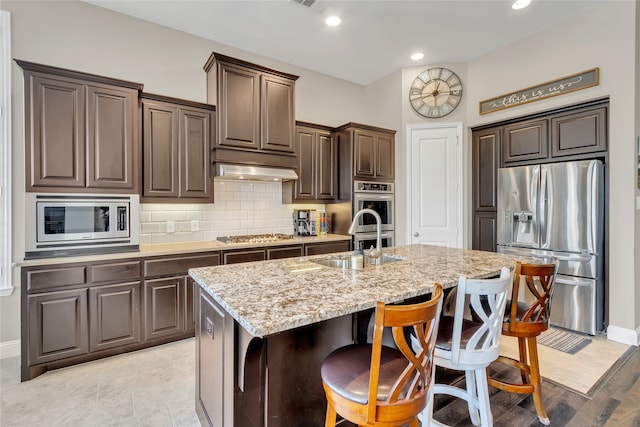 9012 Terrel St, Lantana, TX 76226 - kitchen featuring appliances with stainless steel finishes, a sink, light stone countertops, dark brown cabinets, and a kitchen island with sink