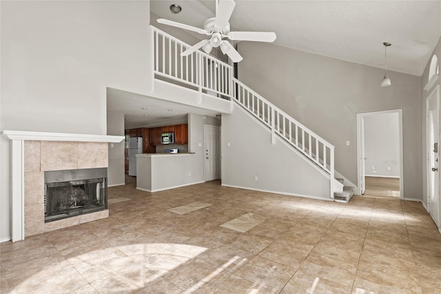 unfurnished living room featuring a ceiling fan, high vaulted ceiling, stairs, a tile fireplace, and baseboards