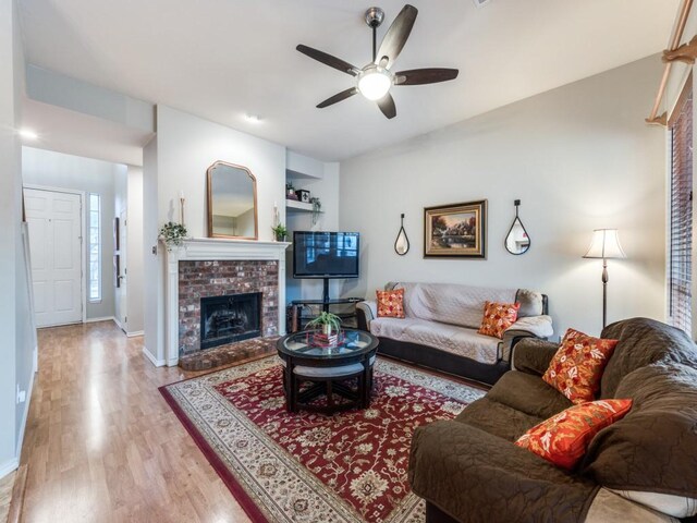 living area featuring wood finished floors, a ceiling fan, baseboards, and a brick fireplace