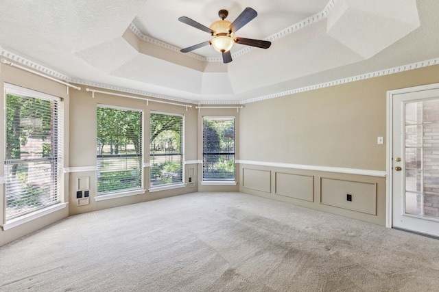 carpeted empty room with a raised ceiling, a ceiling fan, plenty of natural light, a decorative wall, and a wainscoted wall