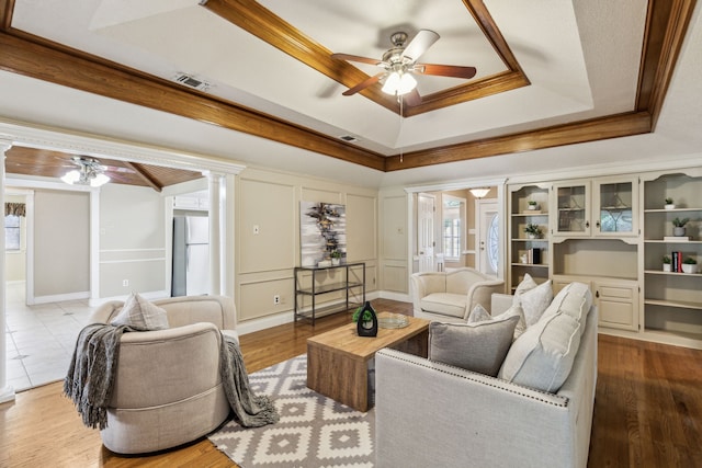 living room featuring ceiling fan, decorative columns, a tray ceiling, wood finished floors, and ornamental molding