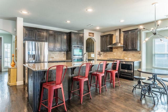 1005 Caudle Ln, Savannah, TX 76227 - kitchen featuring wall chimney range hood, arched walkways, appliances with stainless steel finishes, dark wood-style floors, and a large island with sink