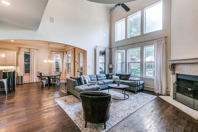 living area with a high ceiling, dark wood-type flooring, a fireplace with raised hearth, arched walkways, and a chandelier