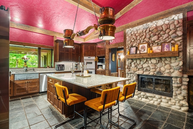 kitchen with dishwasher, stone tile flooring, white double oven, a center island with sink, and a stone fireplace