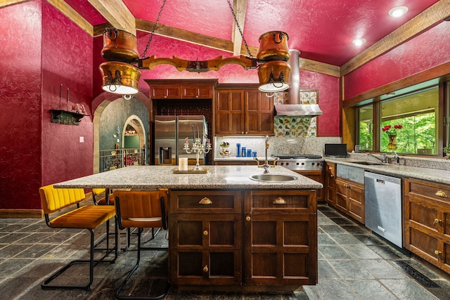kitchen with stainless steel appliances, stone tile floors, a kitchen island with sink, a sink, and a textured wall