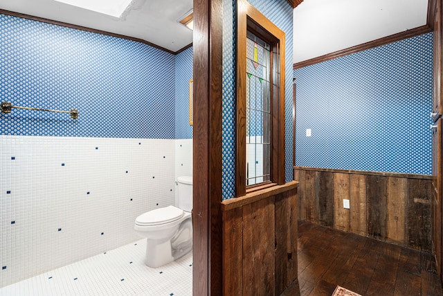 bathroom featuring crown molding, wallpapered walls, toilet, a tile shower, and a wainscoted wall