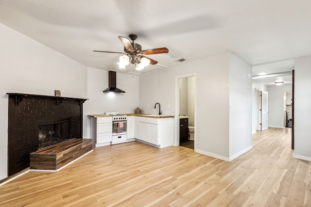3843 Bernal Dr, Dallas, TX 75212 - kitchen featuring range with gas cooktop, wall chimney range hood, ceiling fan, white cabinetry, and a brick fireplace