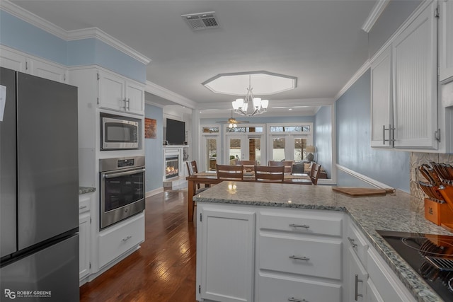 kitchen featuring appliances with stainless steel finishes, a peninsula, a chandelier, white cabinetry, and a warm lit fireplace