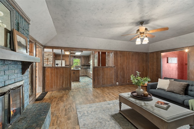 living room featuring lofted ceiling, light wood-style floors, ceiling fan, a brick fireplace, and wooden walls