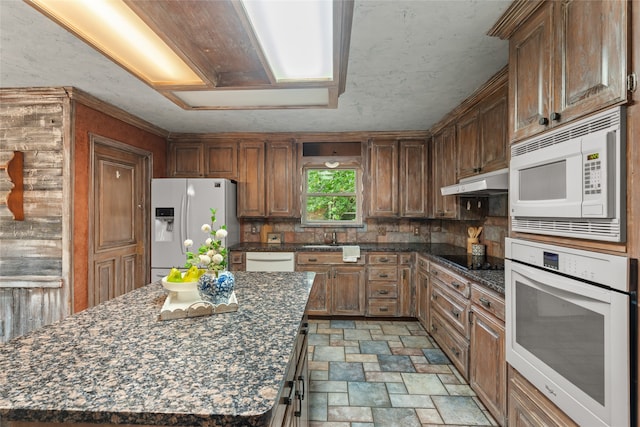 kitchen with white appliances, a sink, stone finish flooring, under cabinet range hood, and backsplash