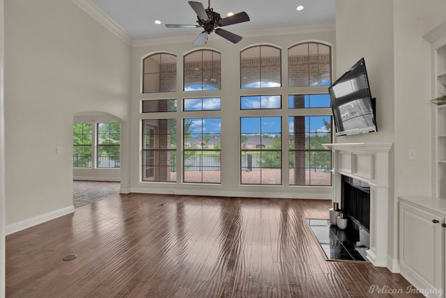 unfurnished living room featuring dark wood-style floors, a fireplace with flush hearth, a ceiling fan, arched walkways, and ornamental molding