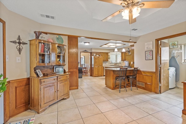 kitchen featuring a peninsula, brown cabinetry, light tile patterned floors, wainscoting, and a ceiling fan