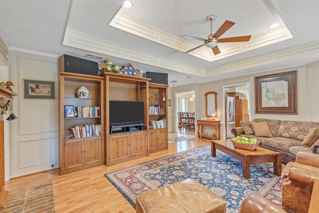 living area featuring ceiling fan, crown molding, light wood-style floors, a textured ceiling, and a tray ceiling