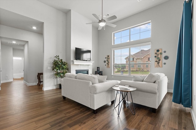 living room featuring a glass covered fireplace, dark wood-style floors, a ceiling fan, baseboards, and a high ceiling