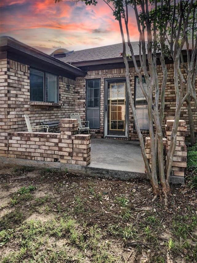 view of exterior entry featuring a patio area, brick siding, and roof with shingles