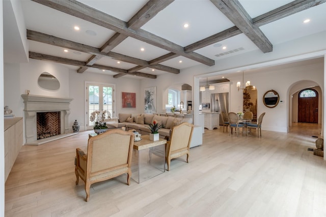 living area with arched walkways, coffered ceiling, light wood-style floors, beamed ceiling, and a fireplace with raised hearth