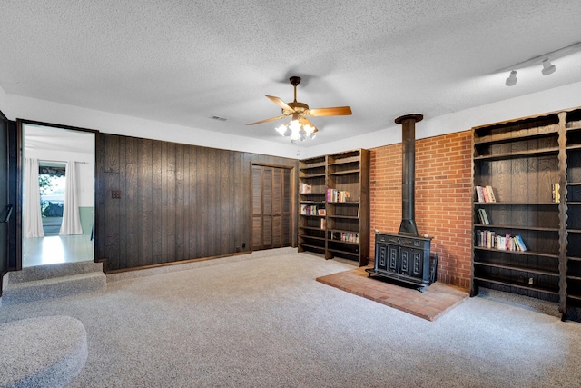 510 Allen Ave, Duncanville, TX 75137 - unfurnished living room with a wood stove, a ceiling fan, wooden walls, a textured ceiling, and carpet flooring