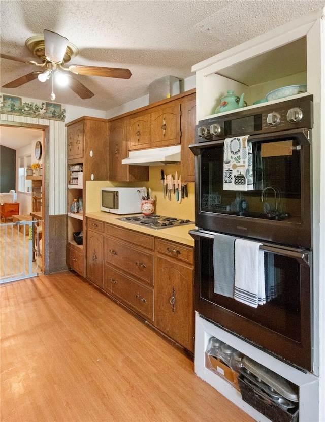 kitchen featuring double oven, brown cabinetry, white microwave, a ceiling fan, and a textured ceiling