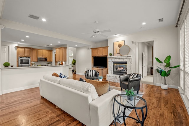 living area featuring a ceiling fan, baseboards, light wood-type flooring, a stone fireplace, and crown molding