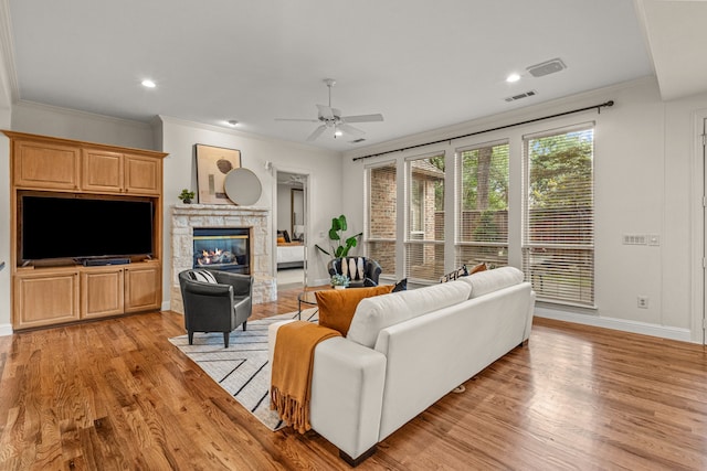 living area featuring a ceiling fan, light wood-style flooring, a stone fireplace, baseboards, and crown molding