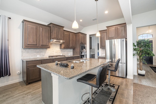 kitchen featuring stainless steel appliances, a sink, light stone counters, baseboards, and a center island with sink