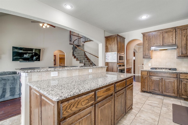 9836 Amaranth Dr, Fort Worth, TX 76177 - kitchen featuring arched walkways, under cabinet range hood, stainless steel appliances, tasteful backsplash, and light stone counters