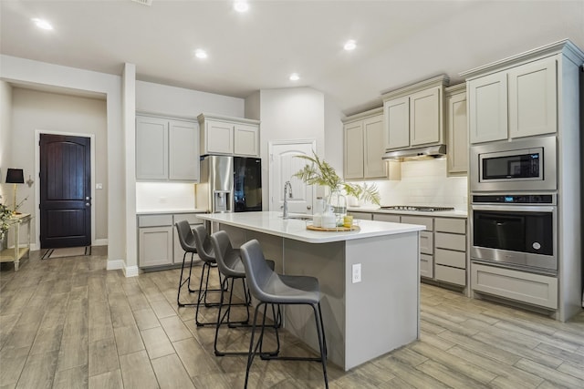 444 Oliver Ct, Argyle, TX 76226 - kitchen featuring appliances with stainless steel finishes, backsplash, a center island with sink, light wood-type flooring, and a breakfast bar