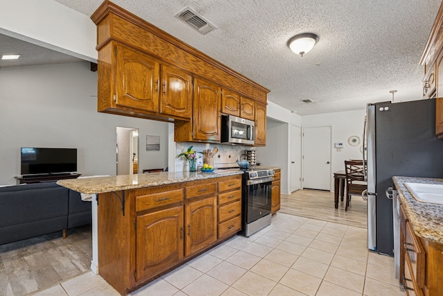 140 Amory Dr, Benbrook, TX 76126 - kitchen featuring stainless steel appliances, a textured ceiling, a peninsula, brown cabinets, and light tile patterned floors