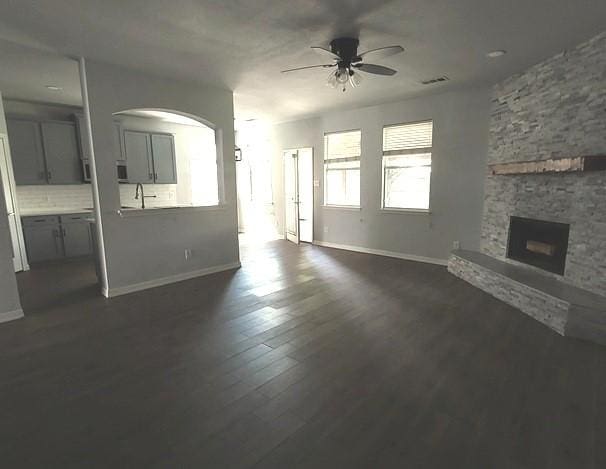 1207 Goldeneye, Aubrey, TX 76227 - unfurnished living room with ceiling fan, a stone fireplace, baseboards, dark wood-type flooring, and arched walkways