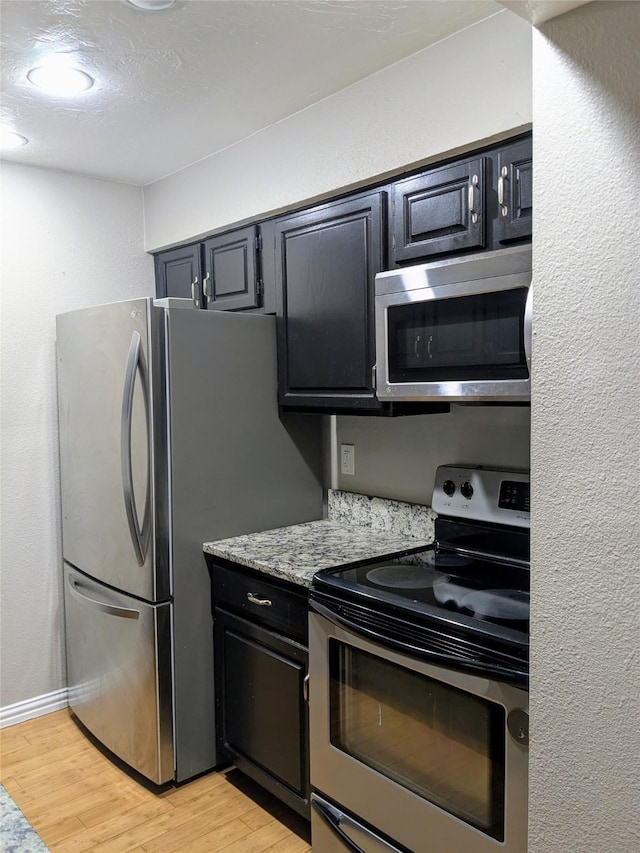 7525 Holly Hill Dr Unit 68, Dallas, TX 75231 - kitchen featuring appliances with stainless steel finishes, light wood finished floors, dark cabinets, a textured wall, and light stone counters