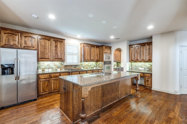 11257 Lamar Ln, Frisco, TX 75033 - kitchen featuring appliances with stainless steel finishes, dark wood-style floors, an island with sink, arched walkways, and light stone counters