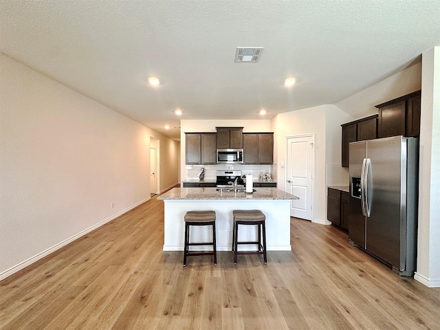 1416 Blossom Court Dr, Princeton, TX 75407 - kitchen featuring appliances with stainless steel finishes, light wood-type flooring, dark brown cabinetry, a center island with sink, and baseboards