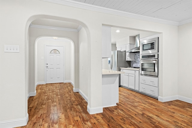 3806 Rainier St, Irving, TX 75062 - kitchen featuring appliances with stainless steel finishes, wall chimney range hood, wood finished floors, white cabinets, and ornamental molding