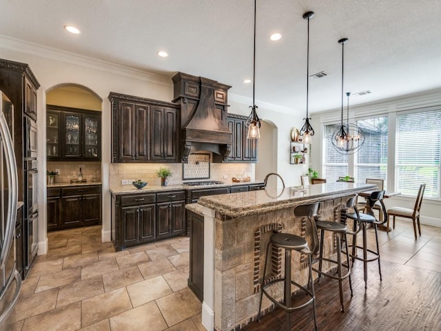 1221 Morris Dr, Keller, TX 76248 - kitchen with arched walkways, custom exhaust hood, decorative backsplash, dark brown cabinets, and a kitchen bar