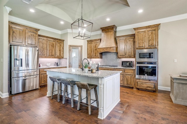 3201 Steamers Ct, Granbury, TX 76049 - kitchen featuring appliances with stainless steel finishes, a kitchen island with sink, brown cabinets, premium range hood, and vaulted ceiling