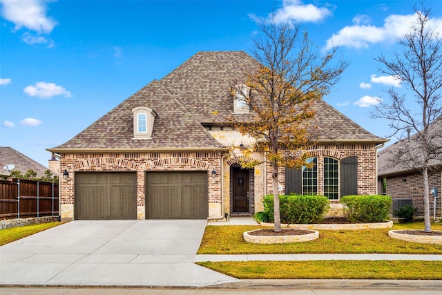 2705 Cumberland, The Colony, TX 75056 - french country home featuring brick siding, concrete driveway, a garage, a shingled roof, and central AC unit