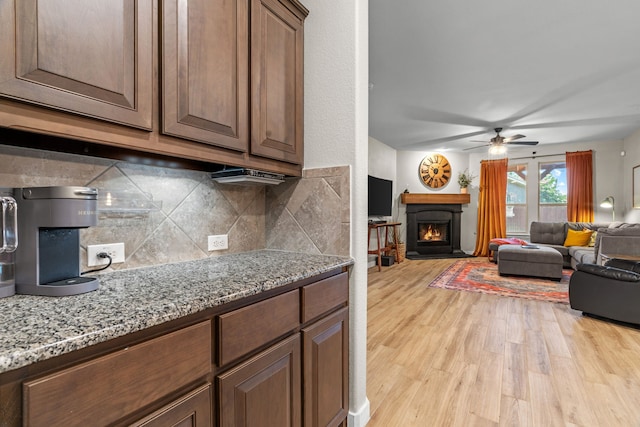15712 Oak Pointe Dr, Fort Worth, TX 76177 - kitchen featuring light wood finished floors, decorative backsplash, a ceiling fan, a warm lit fireplace, and light stone countertops