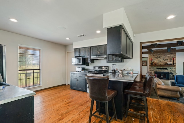 3893 E Fm 906, Powderly, TX 75473 - kitchen with under cabinet range hood, stainless steel appliances, a kitchen breakfast bar, a brick fireplace, and light wood-style floors
