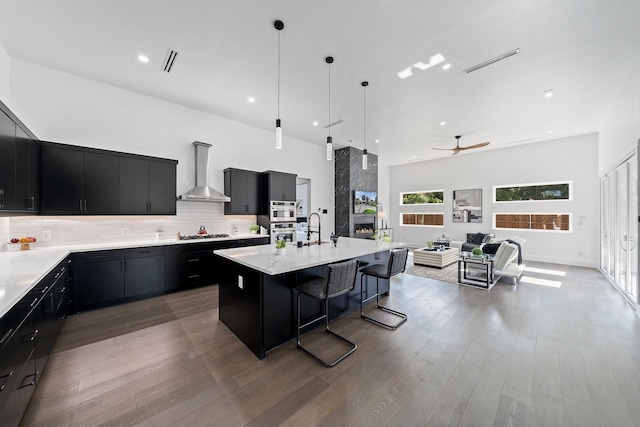 4233 Skillman St, Dallas, TX 75206 - kitchen with dark cabinets, wall chimney range hood, a ceiling fan, a kitchen island with sink, and tasteful backsplash